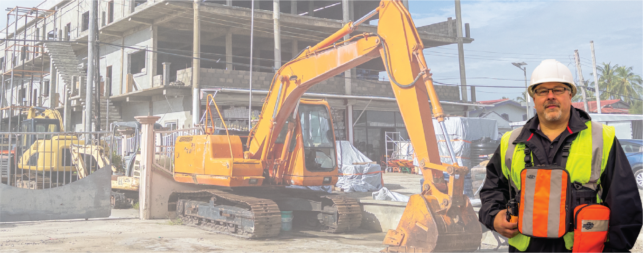 Construction worker wearing a high-visibility vest and a Fitek tablet carry pack, standing in front of an excavator and an active job site with partially built structures.