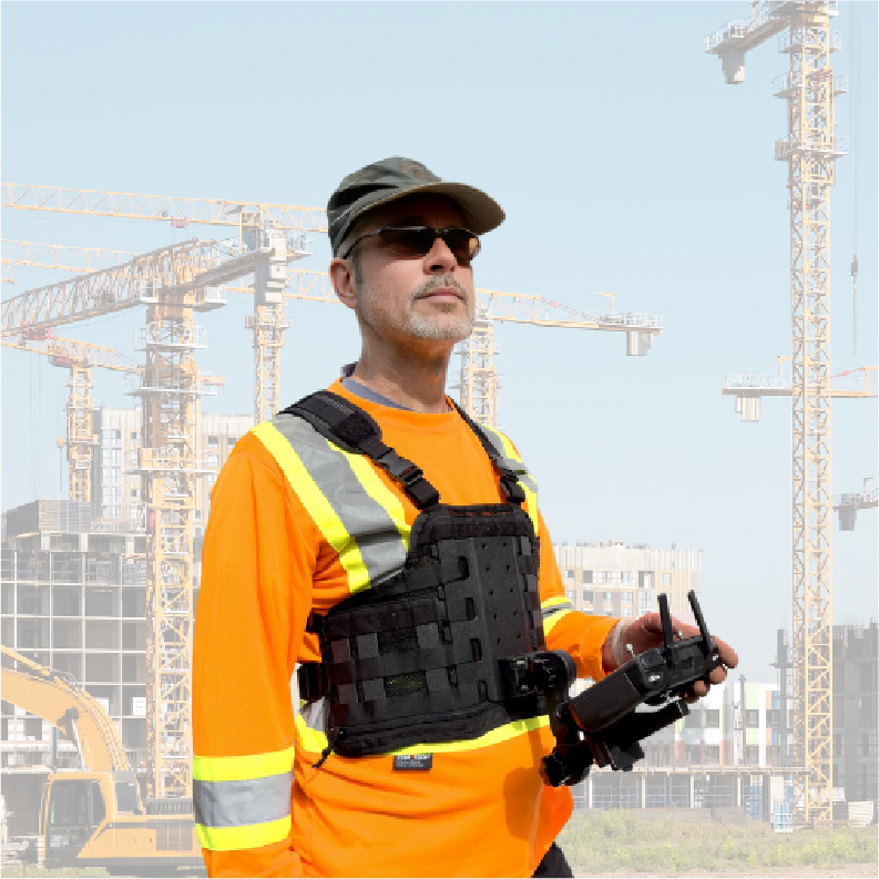 Drone pilot wearing a high-visibility shirt and Fitek drone controller harness, holding a controller in front of an active construction site with cranes and buildings under development