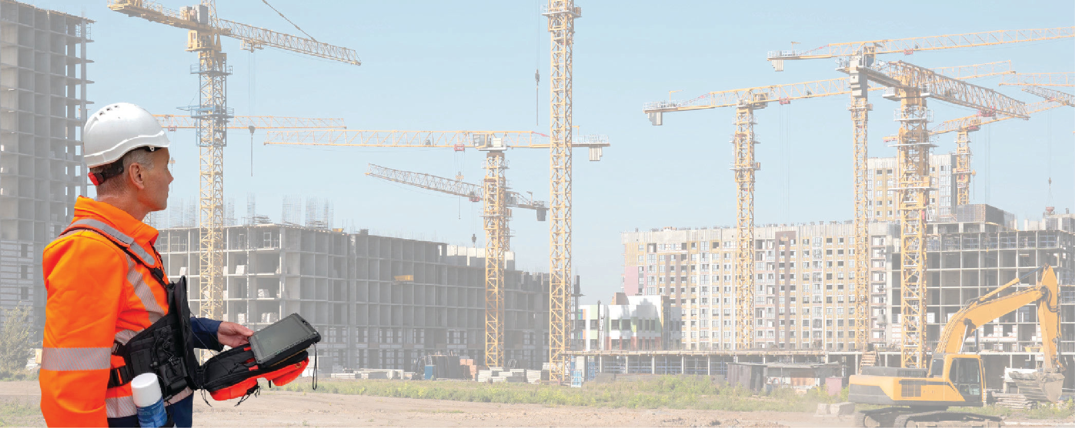 Construction worker wearing a Fitek tablet harness at an active job site, surrounded by cranes and buildings under construction. The tablet is securely mounted on the chest with a fitek chest pack for hands-free access and improved safety.